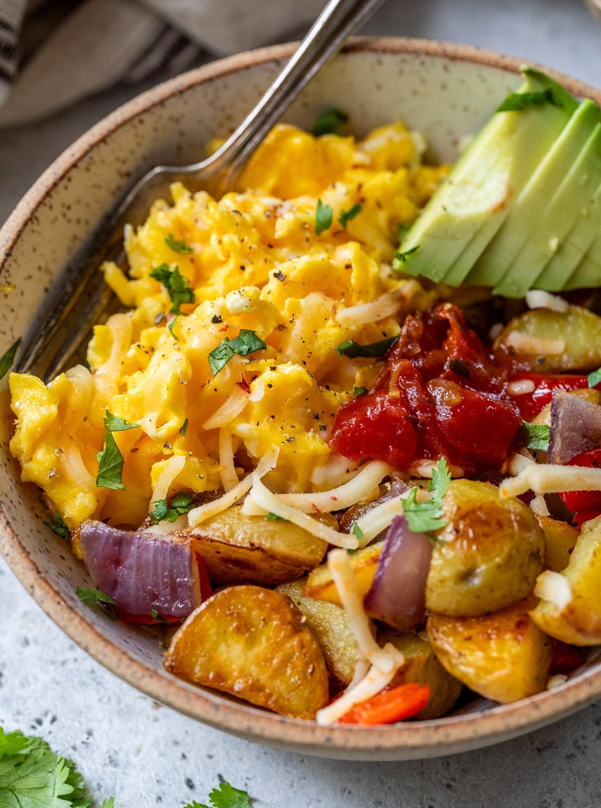 A breakfast bowl of scrambled eggs, roasted potatoes, avocado slices, cheese, salsa, and herbs, with a fork on the side.