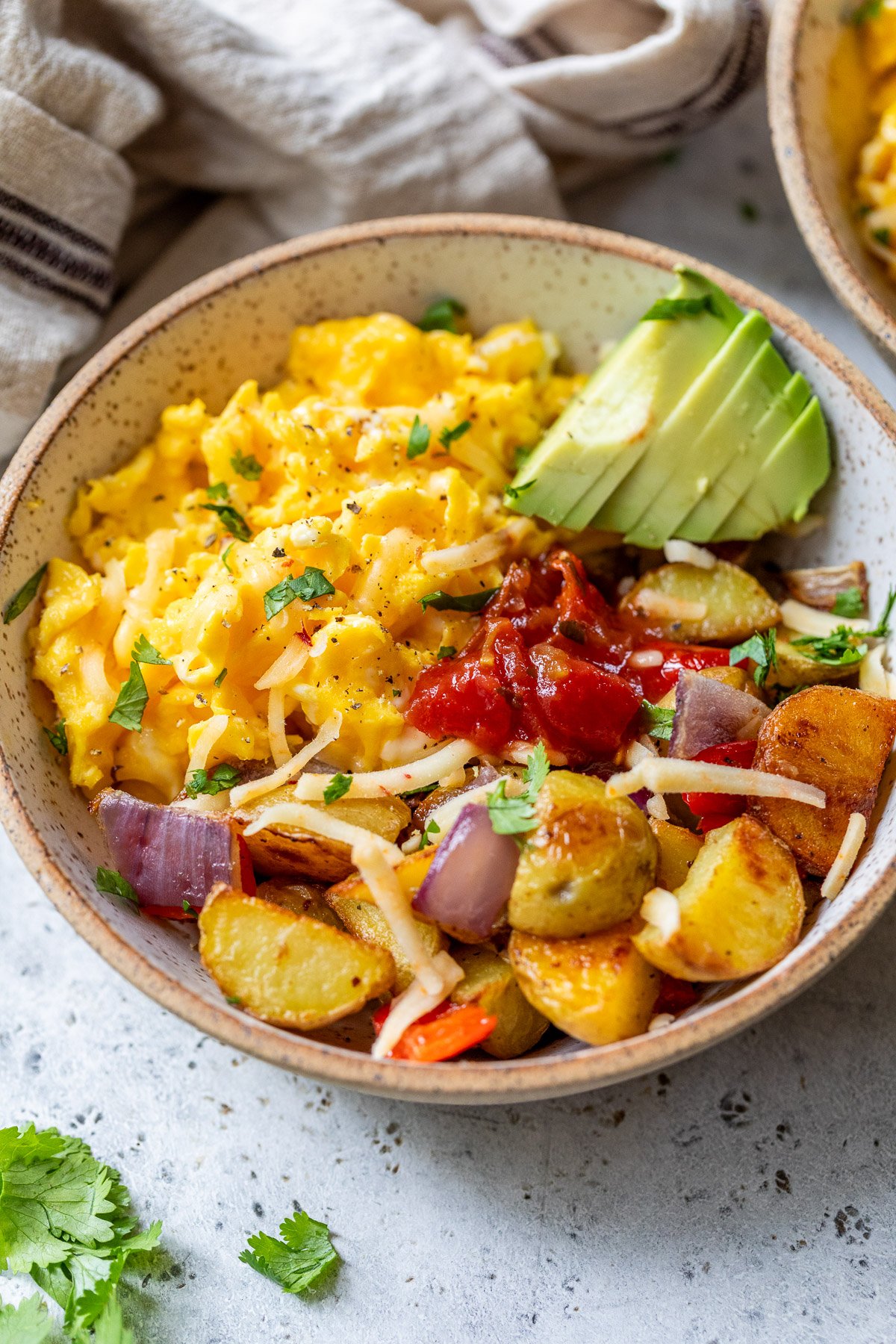 A breakfast bowl with scrambled eggs, avocado slices, salsa, roasted potatoes, onions, and shredded cheese, garnished with cilantro.