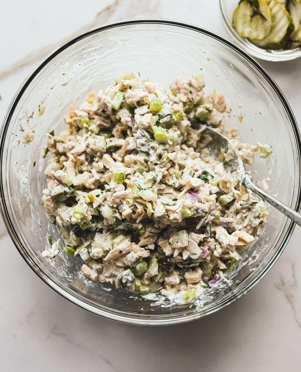A glass bowl filled with creamy chicken salad mixed with chopped celery, red onion, and dill pickles