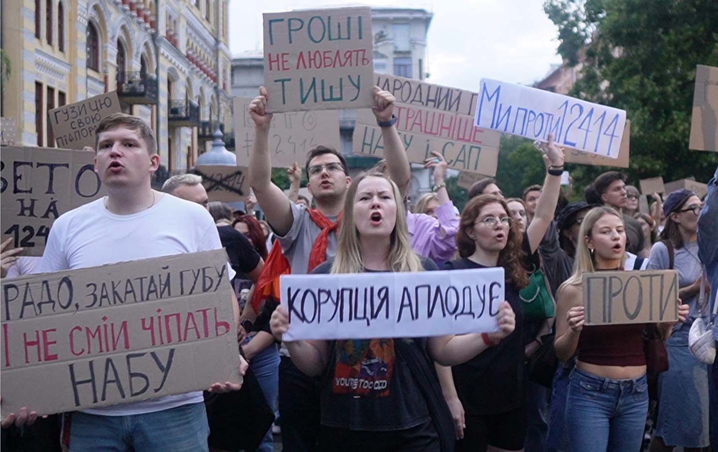 Demonstrators chant outside Kyiv’s Ivan Franko National Drama Theater on July 23, 2025, holding cardboard placards that read “Корупція аплодує” (“Corruption Applauds”) and “Вето на 12444” (“Veto Bill 12444”) to protest new legislation placing Ukraine’s anti‑corruption agencies under presidential control.