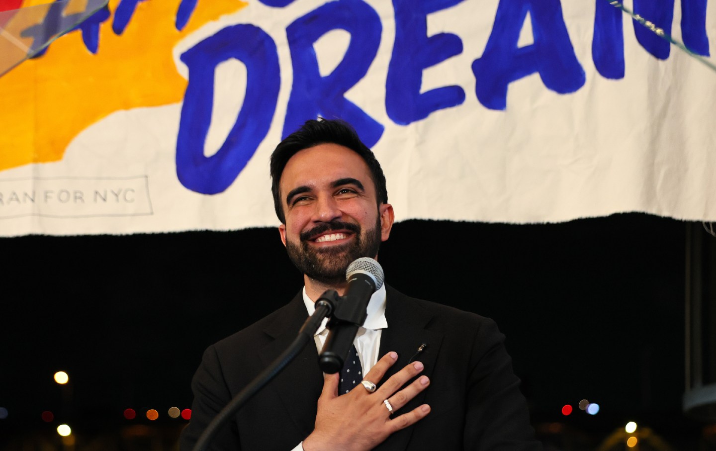 Zohran Mamdani smiles in front of a microphone with his hand on his chest.