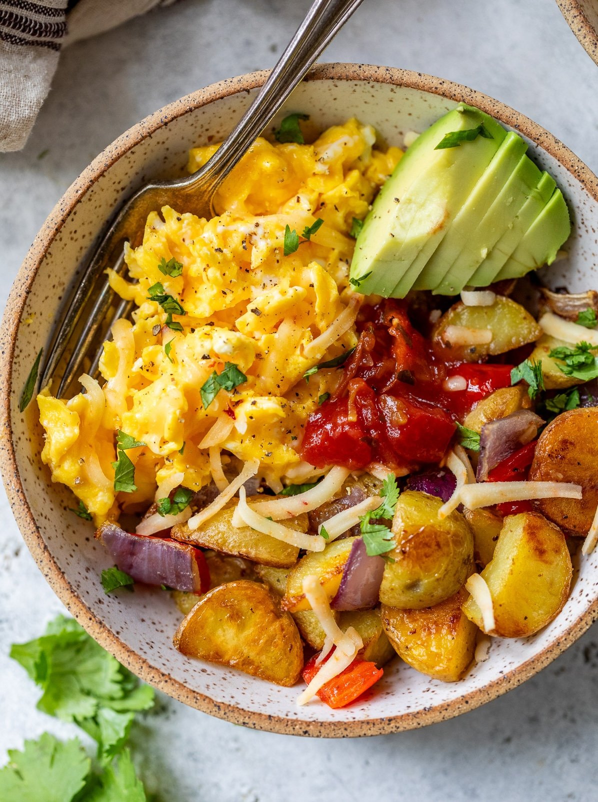 A breakfast bowl with scrambled eggs, sliced avocado, roasted potatoes, salsa, shredded cheese, and herbs.