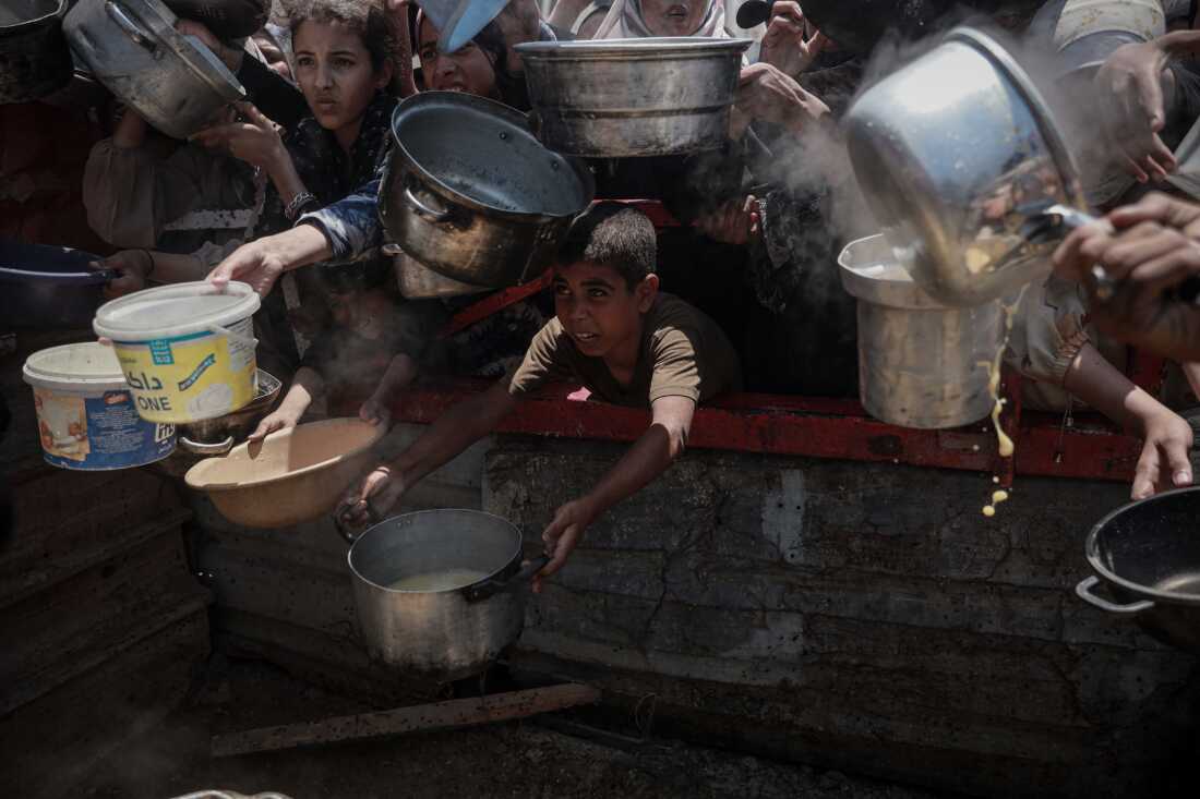 In this photo, Palestinian people, including children, are holding out metal pots and plastic tubs as they wait for food aid in Gaza City on July 27, 2025.