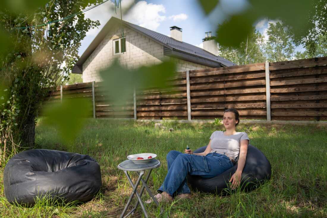 In this photo, Liia Kazakova sits outdoors on a beanbag chair in a grassy area. A brown wooden fence rises in the background.