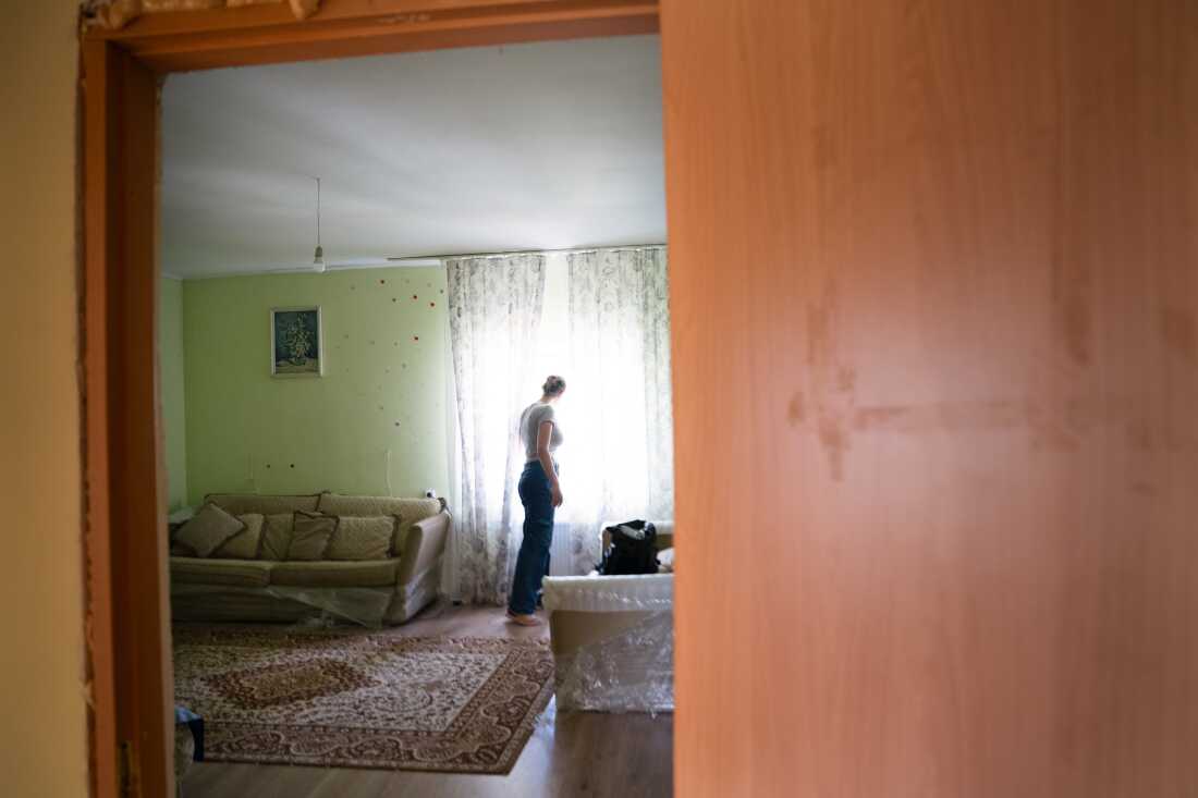 In this photo, Liia Kazakova is standing in a living room with a sofa and rug in it and is looking out through the parted curtains of a window.
