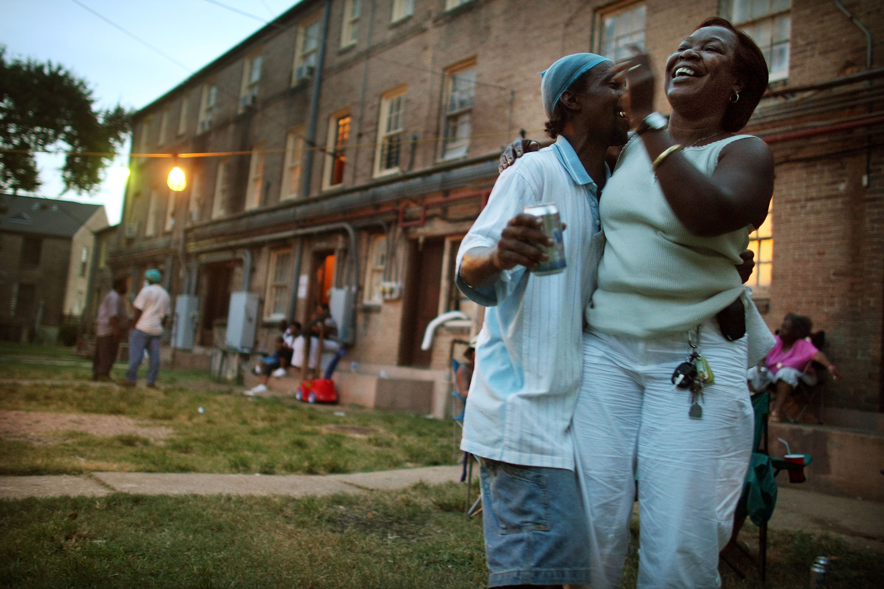 A photo of a man and woman dancing in a blur in the dimly lit courtyard of an apartment building. Other people are also hanging out in the background.