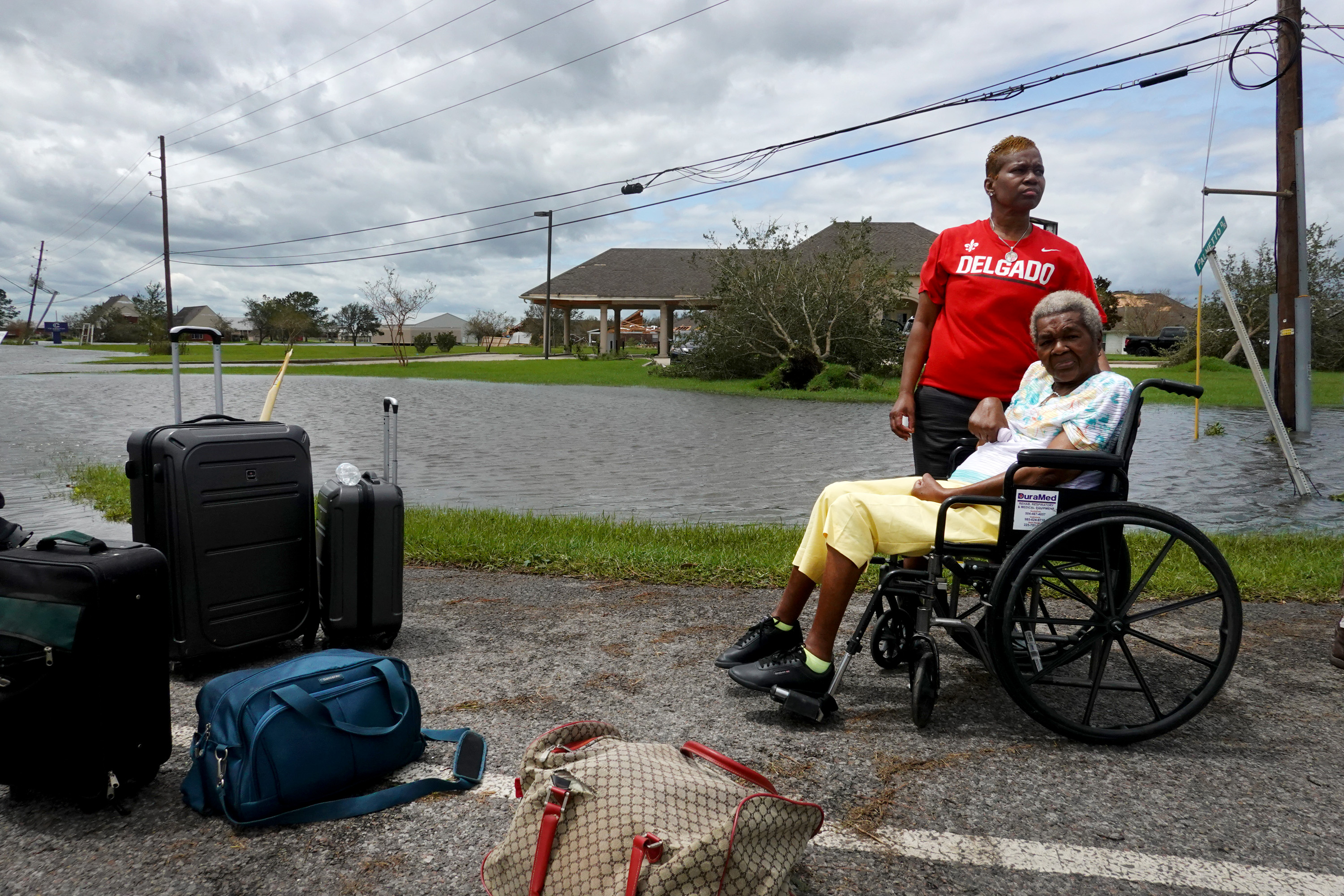 A photo of an elderly woman in a wheelchair and a middle-aged woman standing beside her. Beside them are bags of luggage. In the background is a neighborhood under water.