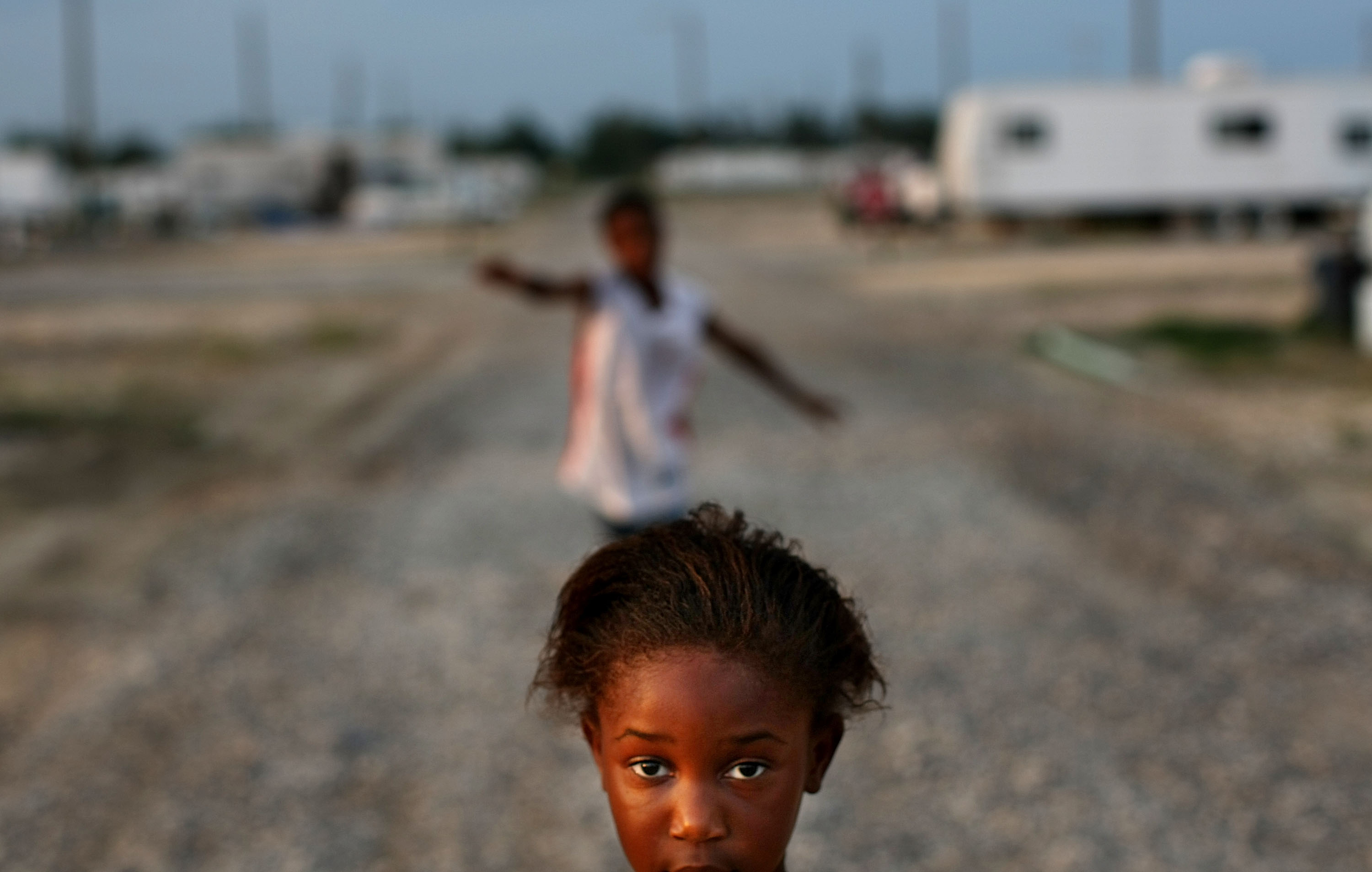 A close-up photo of a young child looking into the lens of a camera. In the background out of focus is a person with splayed arms splayed and a park of trailers.