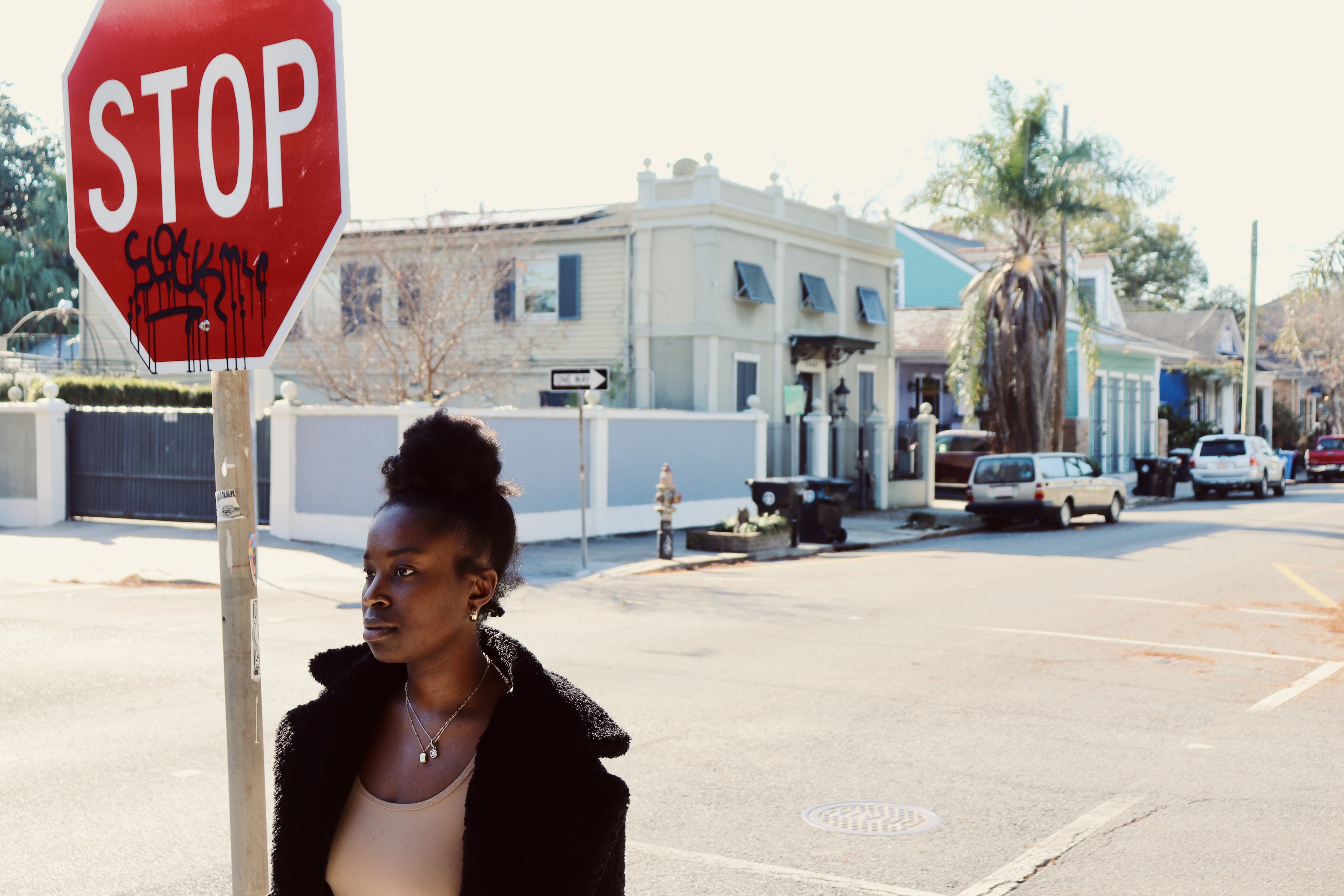 A woman stands under a stop sign on the sidewalk at an intersection of streets.