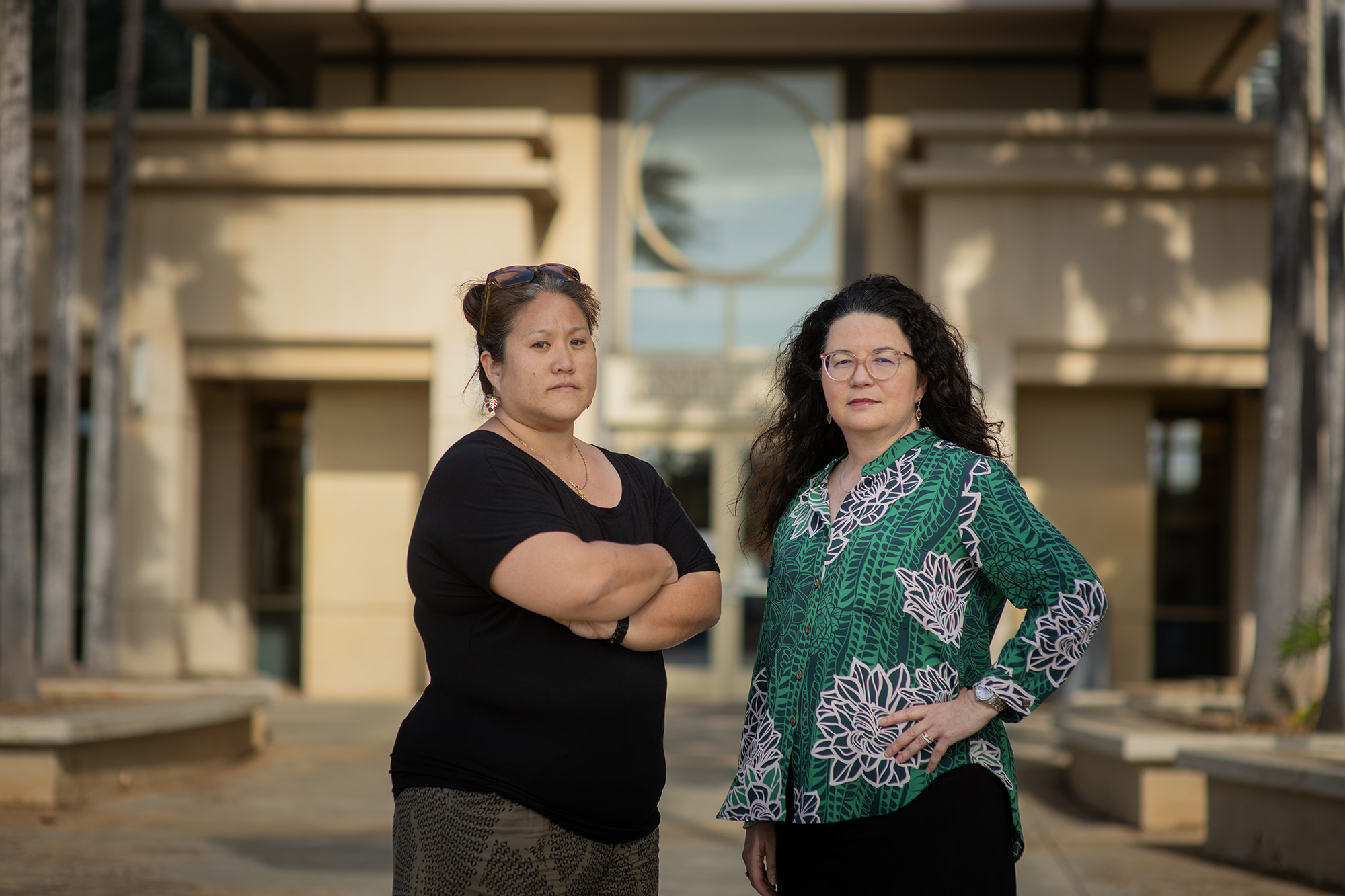 Two women pose in front of a courthouse on Oahu. Tia Ikeno wears a black top and stands with arms crossed, looking determined. Her hair is pulled back in a bun. Judge Dyan Medeiros has a green top and glasses, with long dark wavy hair