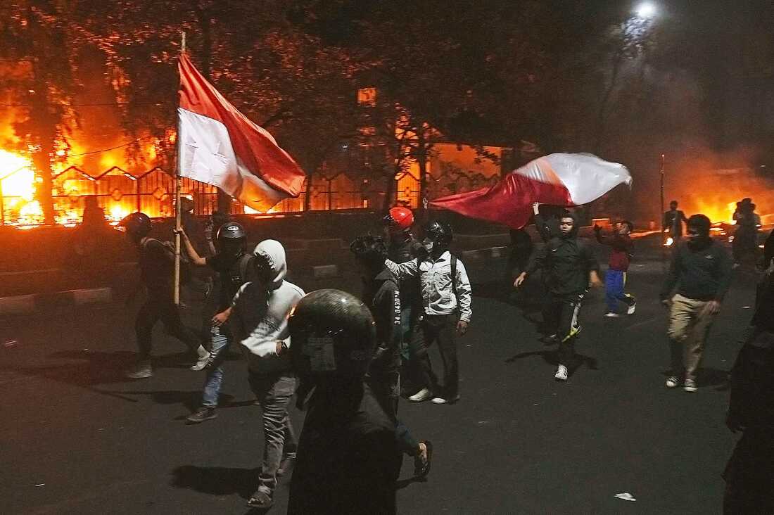 Protesters walk by as the local parliament building is engulfed in flames during a protest following the death of a delivery rider in clashes between riot police and students protesting against lawmakers