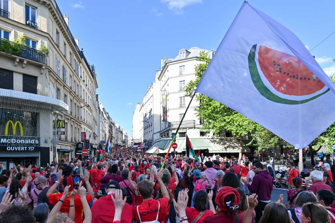 A white flag with a large watermelon slice on it is waved during the Red Line for Gaza protest in Paris on July 8. Most of the protesters are wearing red shirts, and they're walking down a street lined by multistory buildings.