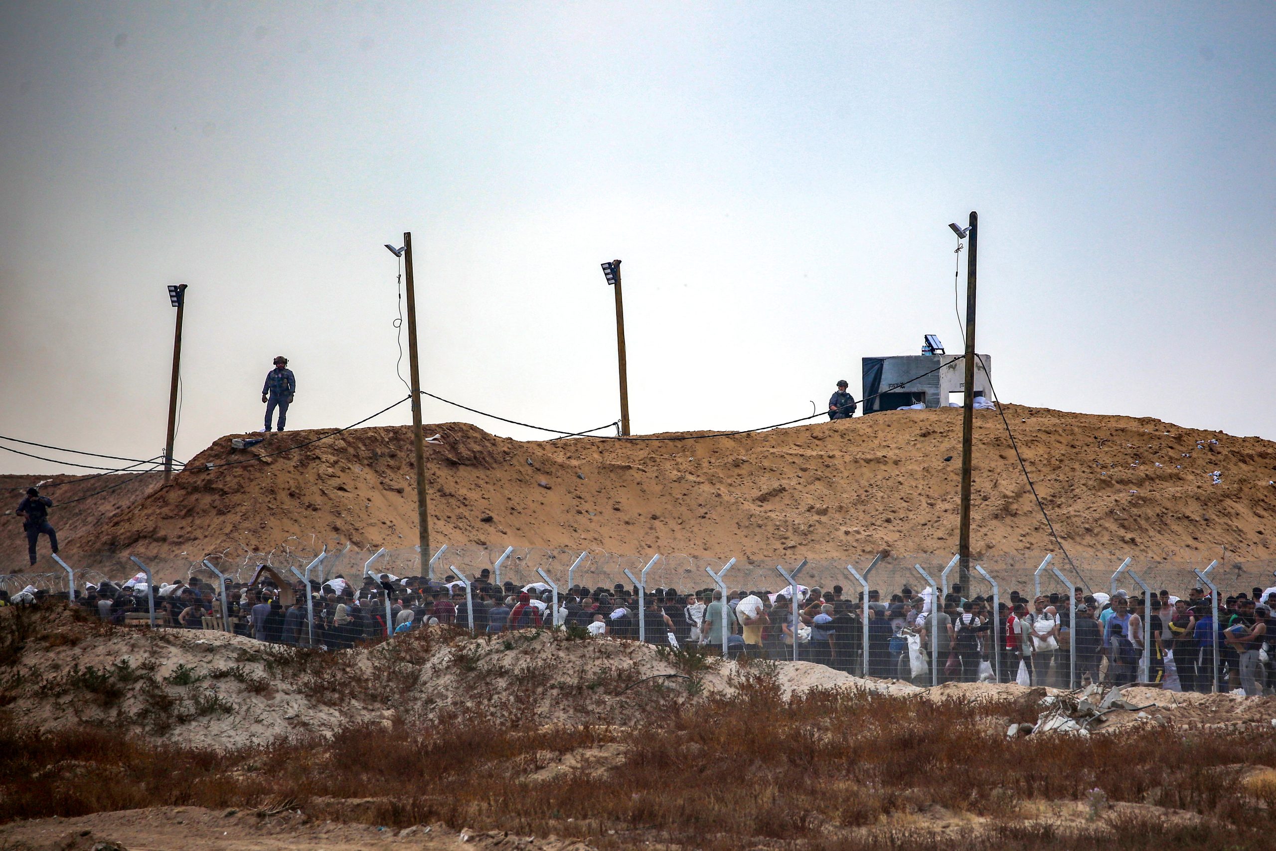 Three security guards are silhouetted on a dirt hill above a fenced-in corridor, where a large crowd of Palestinians line up to collect boxes of aid.