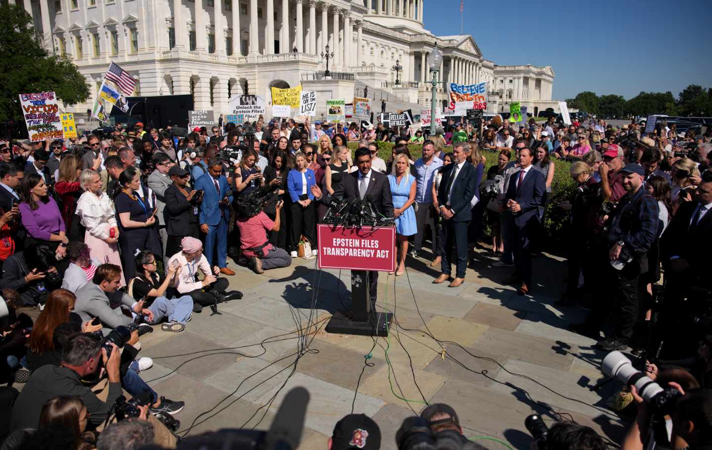 Representative Ro Khanna at the center of a wide circle of people, at a podium, near the Capitol, with a sign reading