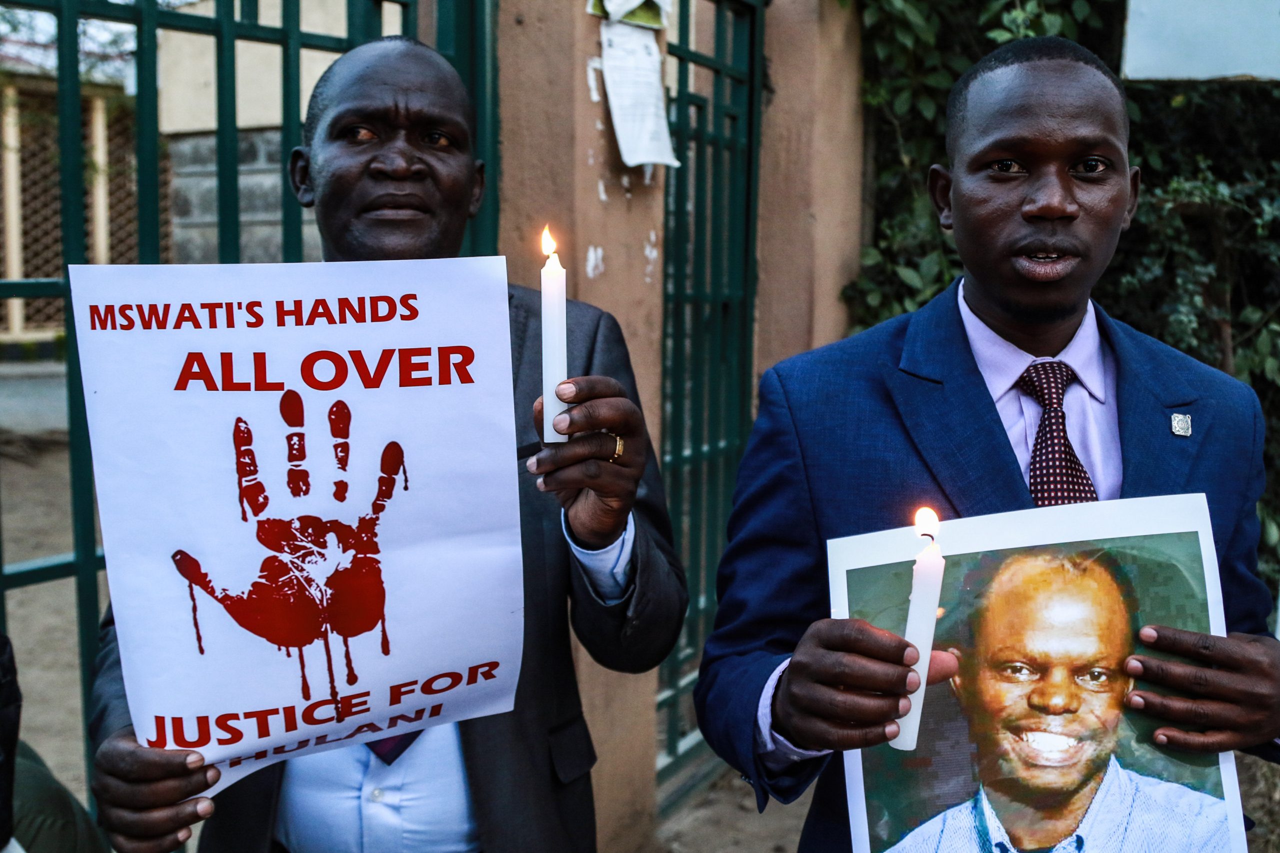 Two men stand in front of a gate holding candles, one holding a signs with a bloody hand print, the other holding a photo of a smiling man.