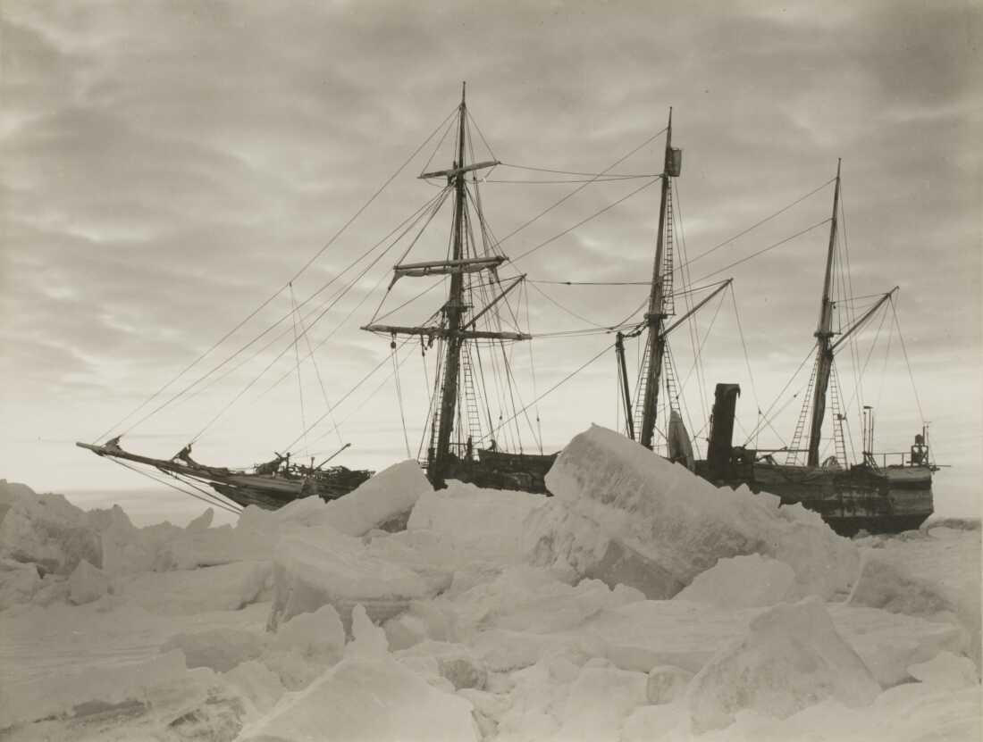 This black-and-white photo shows the ship Endurance in the winter of 1915. Large pieces of sea ice are in the foreground, with the three-masted ship behind the ice.