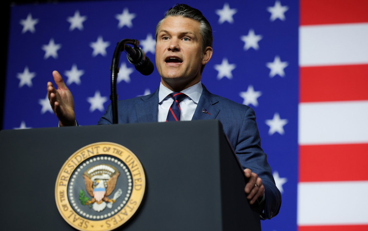 Secretary of Defense Pete Hegseth at a podium gesticulating in front of a large American flag.