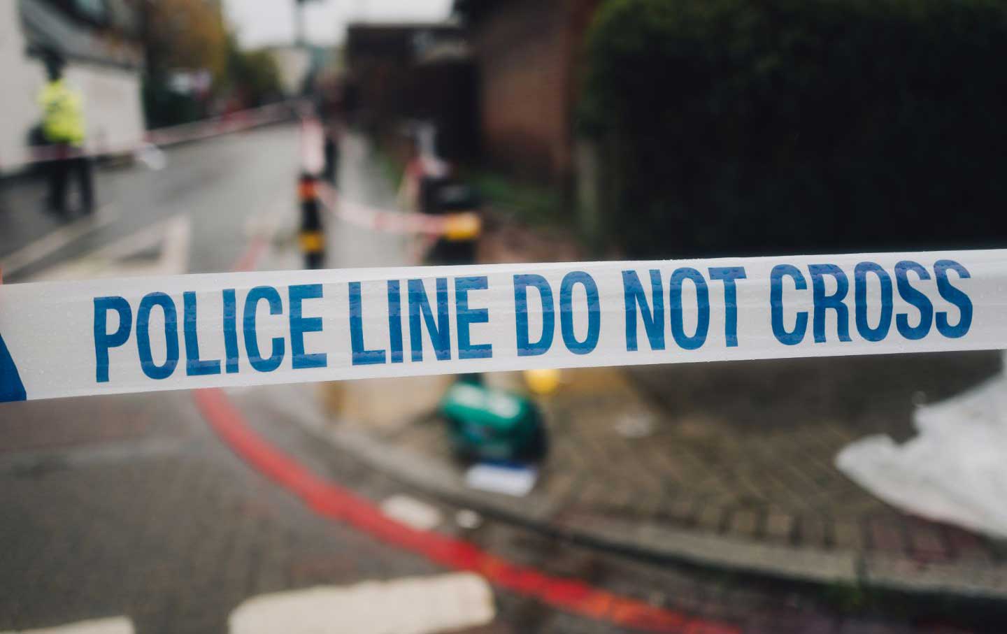 A stock photo of caution tape reading “Police Line Do Not Cross” across a suburban road.